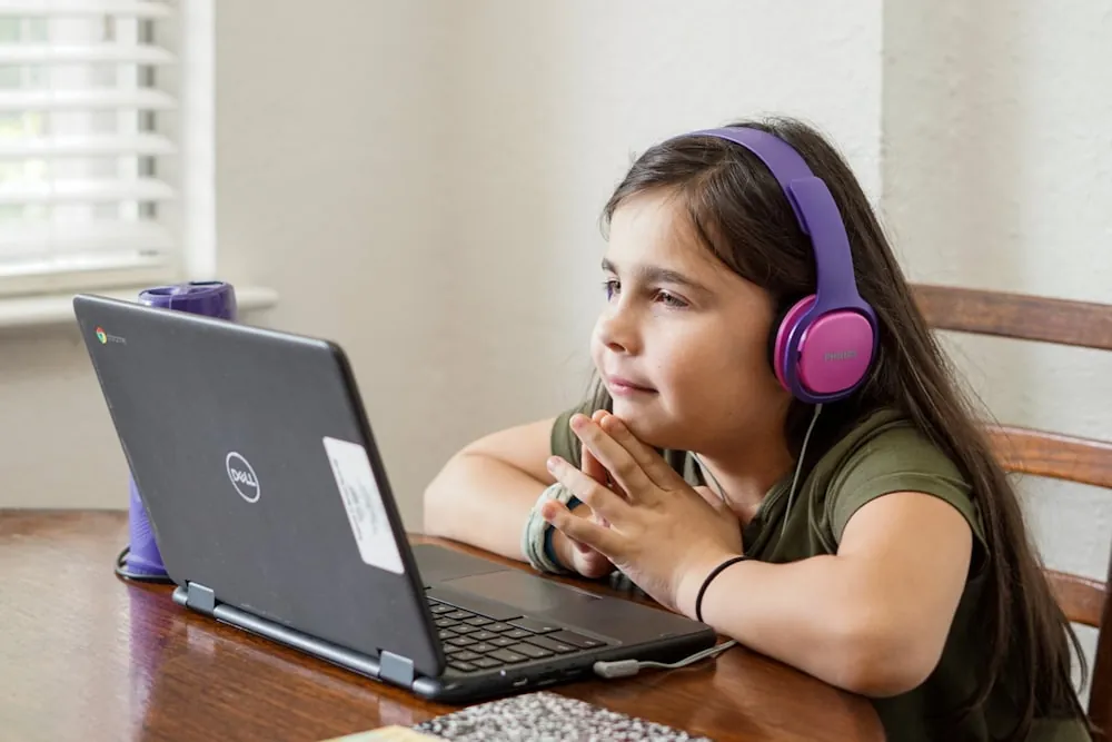 A smiling girl in purple headphones, hands clasped, follows a live class on her laptop at home