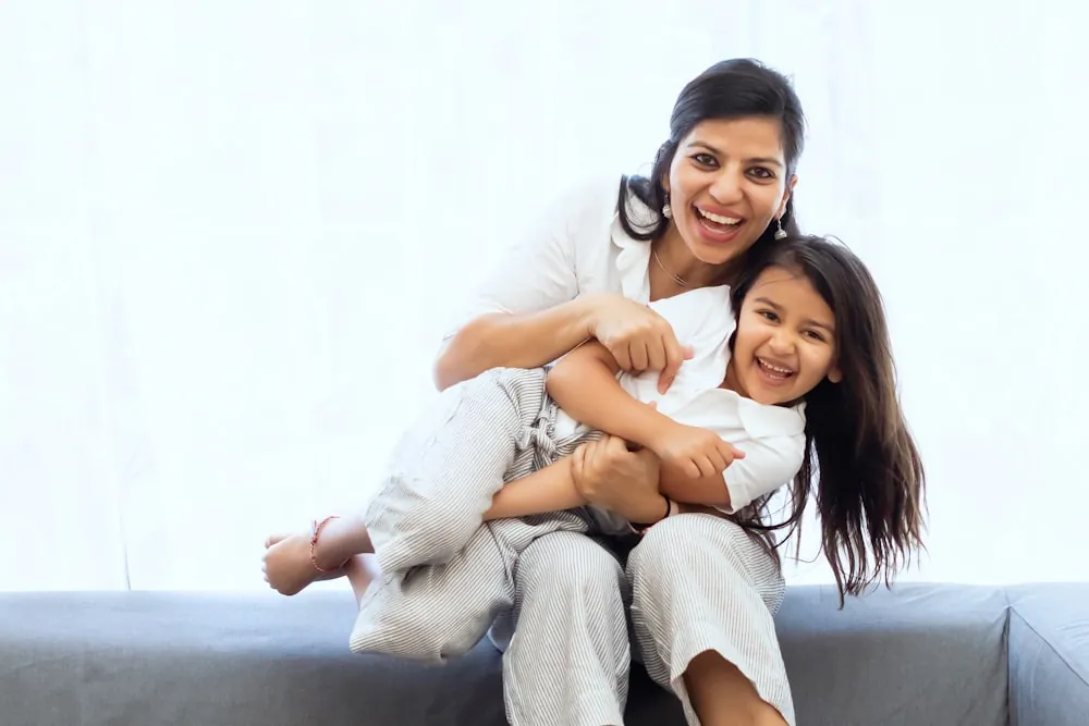 A laughing mother hugs her young daughter on a sofa