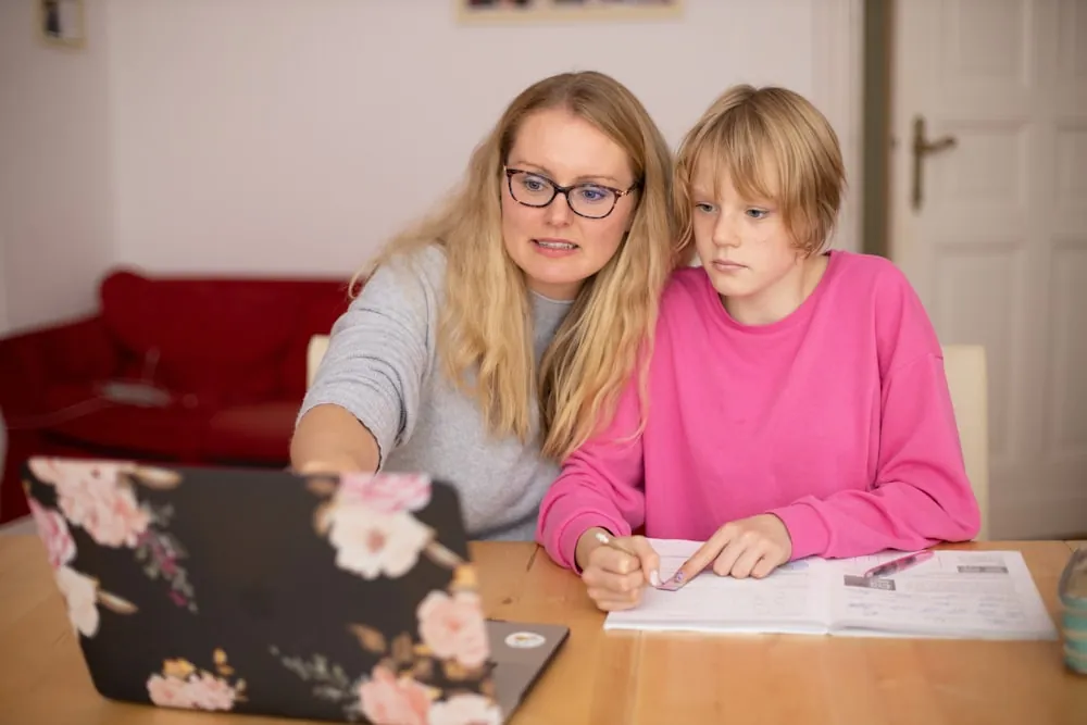 A woman in glasses sits beside a girl in a pink jumper, guiding her through a workbook lesson next to a laptop