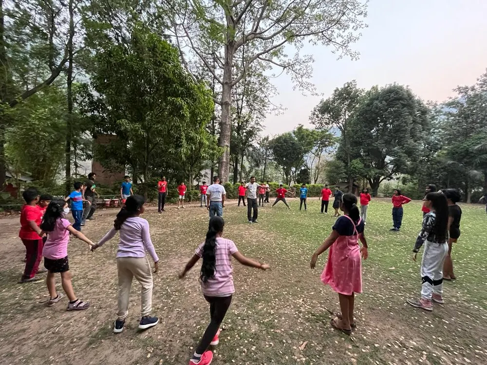Learners playing a circle game on a lawn under tall trees